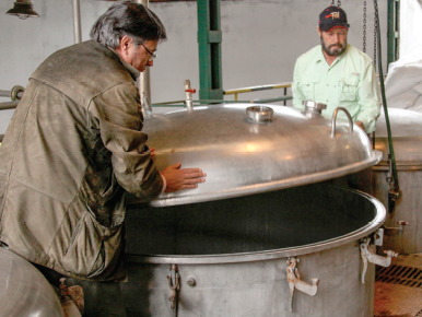 Two men lowering the large metal lid of a distilling chamber.
